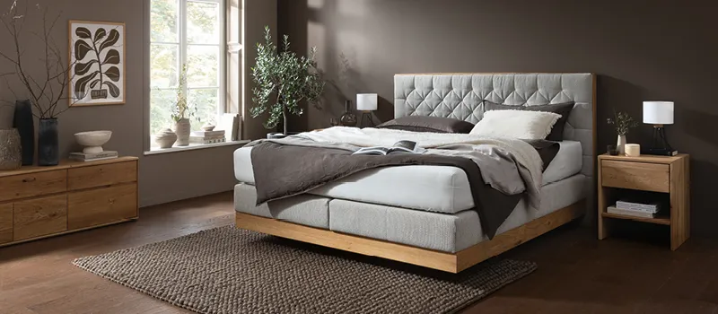 Helles Schlafzimmer mit komfortablem Boxspringbett und gepolstertem Kopfteil, Holzrahmen und passendem Nachttisch, ergänzt durch natürliche Dekoration und harmonische Farbgestaltung für gemütliche Schlafatmosphäre.